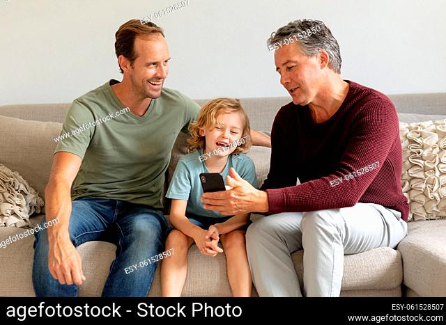 Senior caucasian grandfather on couch looking at smartphone with adult son and grandson laughing
