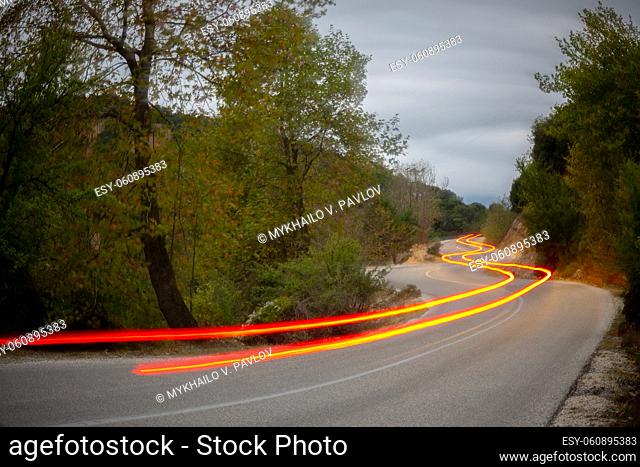 Rear lights of the car illuminate an empty road in the evening summer forest. Long winding trails