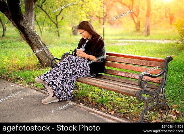 Woman of Asian ethnicity in beautiful long dress sits at park on old rusty bench and reads book using her gadget or checks social networks
