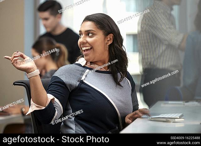 Young woman at computer in office looking over shoulder pointing, co-workers having discussion around dry erase board in background