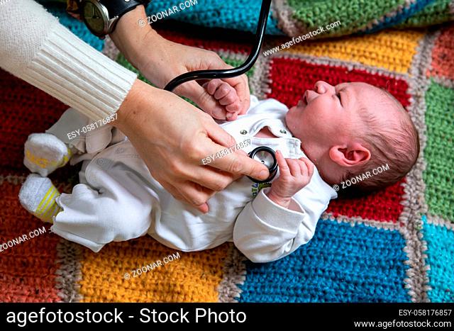 Selective focus of hands of young female doctor holding newborn crying baby hands on knitted bed and checking health using stethoscope
