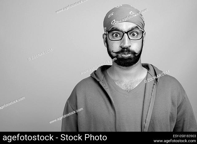 Studio shot of young handsome bearded Indian man wearing eyeglasses against gray background in black and white