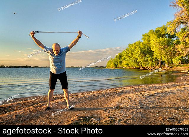 senior man is stretching and exercising with a jump rope at sunrise on a lake beach - Boyd Lake State Park in northern Colorado