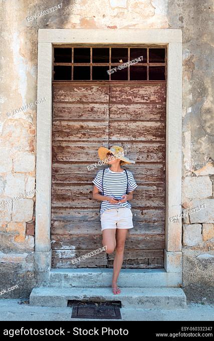 Beautiful young female tourist woman standing in front of vinatage wooden door and textured stone wall at old Mediterranean town, smiling, holding