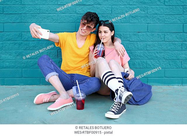 Young couple with soft drinks taking selfie with smartphone in front of blue brick wall