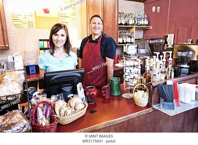 Caucasian woman and man, business owners of a coffee shop, at the counter. Displays of fresh baked goods