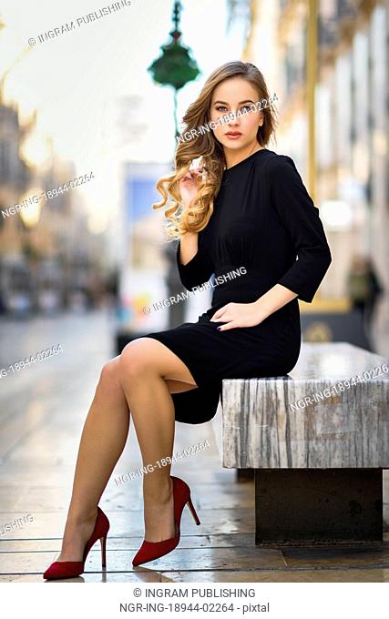 Blonde woman in urban background. Beautiful young girl wearing black elegant dress and red high heels standing in the street