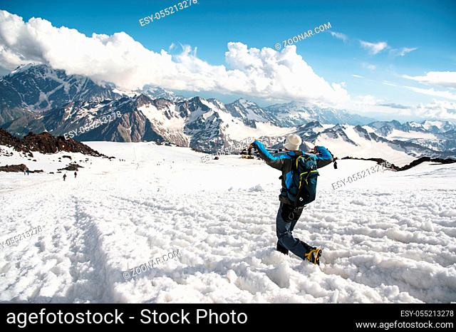 Tired traveler in sunglasses and with a backpack on which the ice-ax is weighing down down from a snowy peak against the background of snow-capped mountains and...