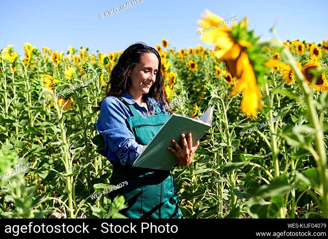 Mature female farm worker writing in book while standing amidst sunflowers