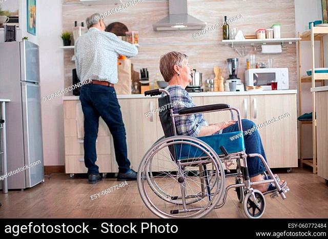 Disabled senior woman sitting in wheelchair in kitchen looking through window. Living with handicapped person. Husband helping wife with disability