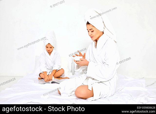 happy caucasian mother in white bathrobes and towels on head apply cream on hands and her daughter reading magazine on background