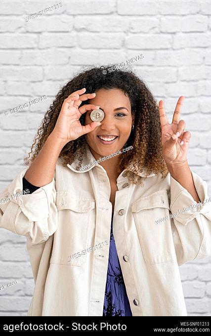 Smiling young woman showing peace sign while covering eye with bitcoin in front of wall