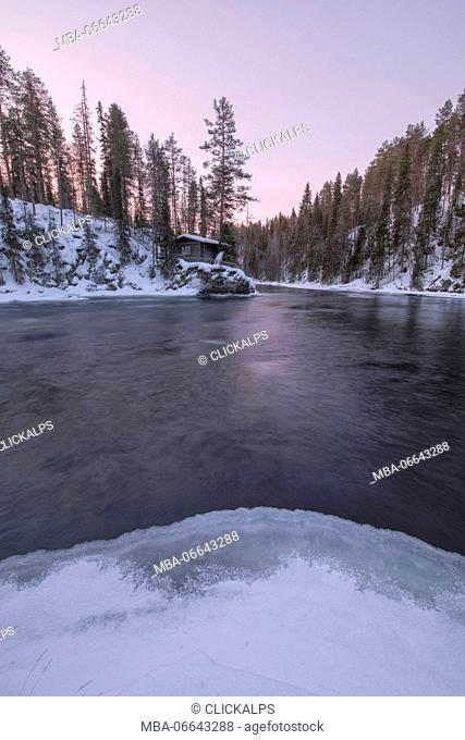 A wooden hut surrounded by the frozen river and snowy woods at dusk Juuma Myllykoski Lapland region Finland Europe