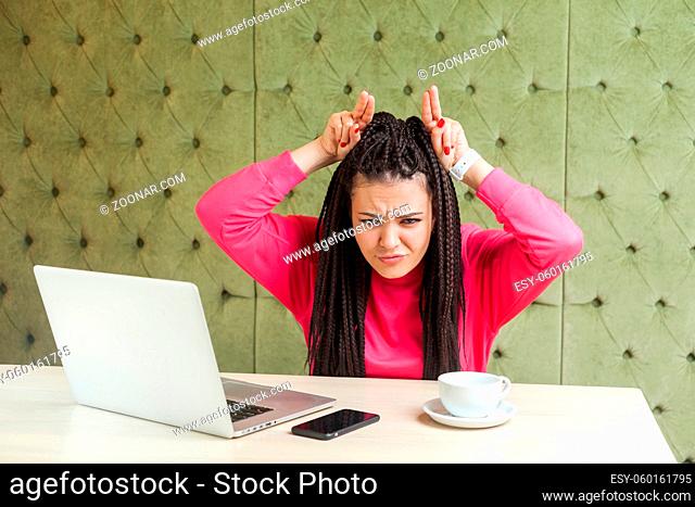 Portrait of aggressive angry young girl freelancer with black dreadlocks hairstyle in pink blouse are sitting in cafe and having bad mood are demonstrate bully...