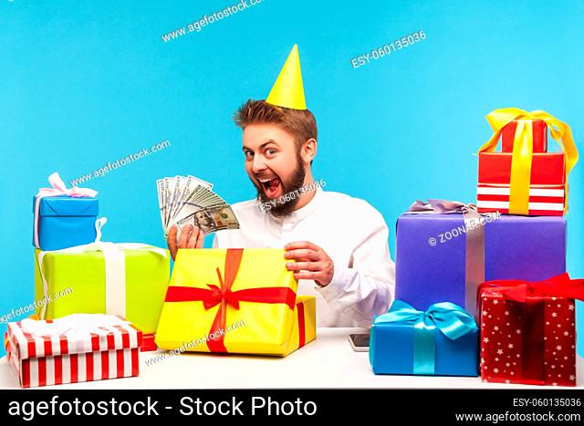 Happy excited man in party cap opening one of many gift boxes and showing lot of dollar cash, satisfied with generous cash birthday present, bonuses