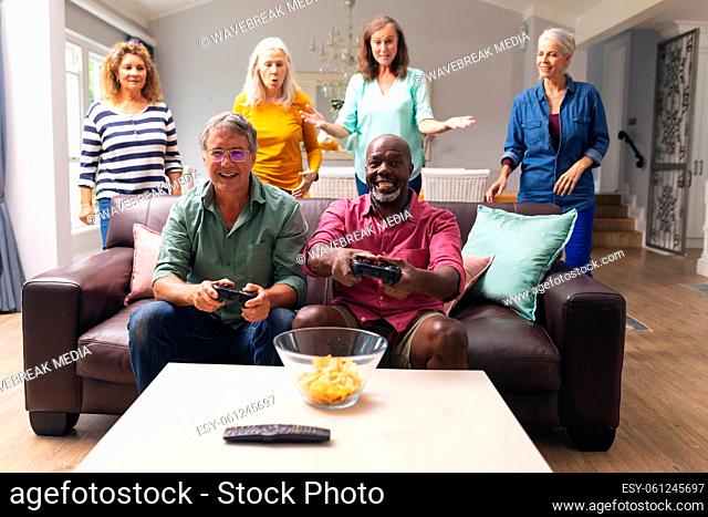 Happy multiracial senior male friends playing video game while women in background at home