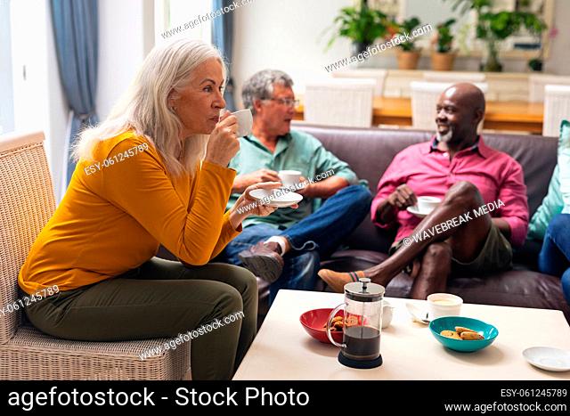 Senior woman drinking coffee while multiracial friends talking in background at home