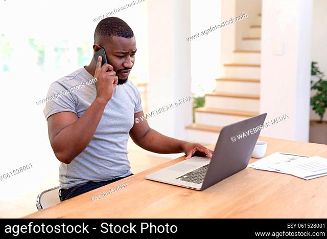 African american man working from home sitting at table using laptop and talking on smartphone