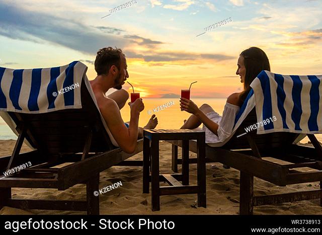 Rear view of a young romantic couple in love sitting on wooden chairs, while drinking cocktails on a tropical beach at sunset during vacation or honeymoon in...