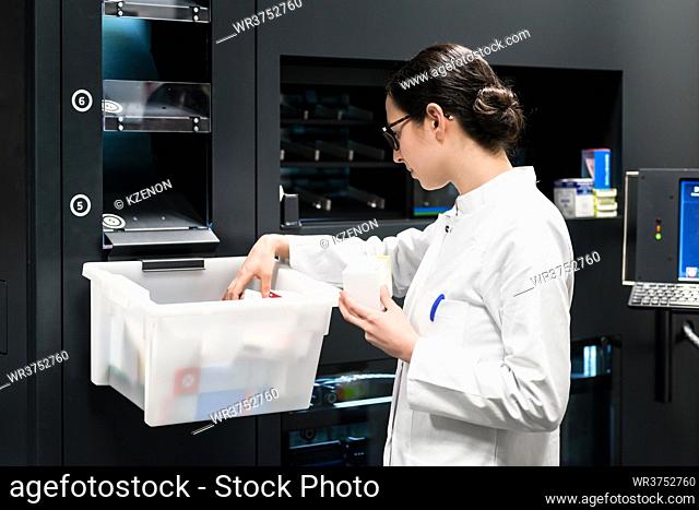 Side view of a dedicated female pharmacist choosing the best medicines from a plastic box during work in a contemporary drugstore with modern equipment