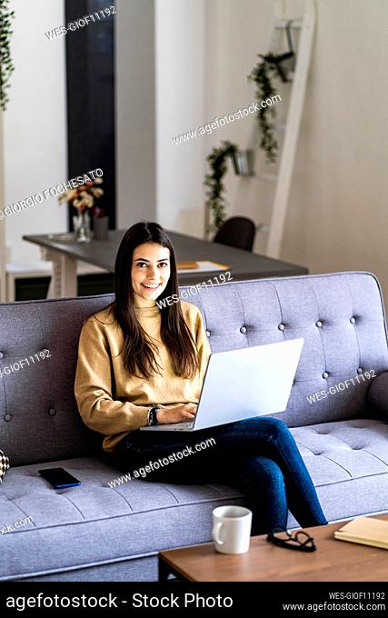 Smiling woman using laptop while sitting with legs crossed at knee on sofa at home
