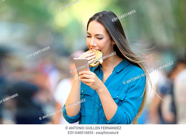 Happy woman eating a burger holding a smart phone on the street