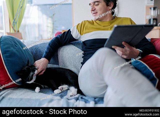 Smiling man with digital tablet stroking cat while sitting on sofa in living room