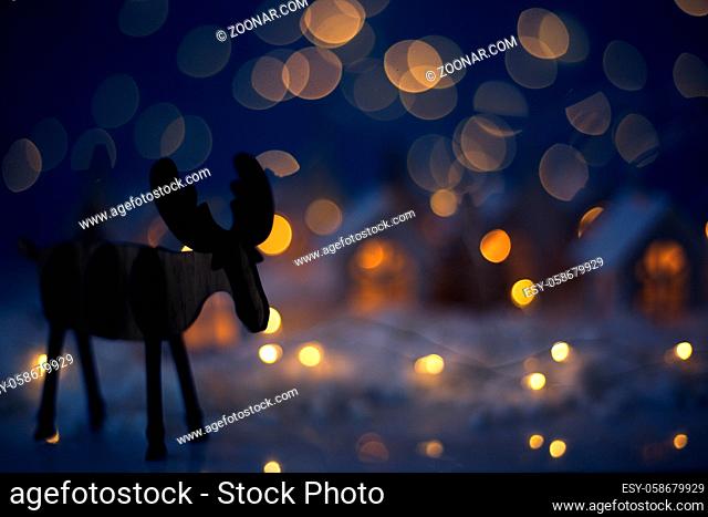 Christmas wooden reindeer over small glowing toy houses village garland over bokeh lights background with copy space for text