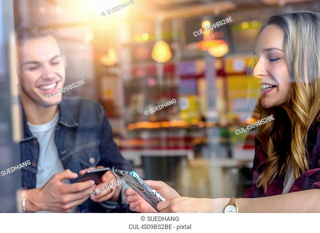 Male and female students looking at smartphones in cafe window seat, view through window