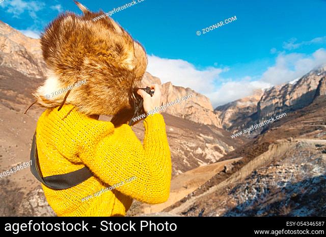 Girl tourist photographer in a fur hat and yellow sweater in the mountains takes pictures on her digital camera. The concept of photography in travel