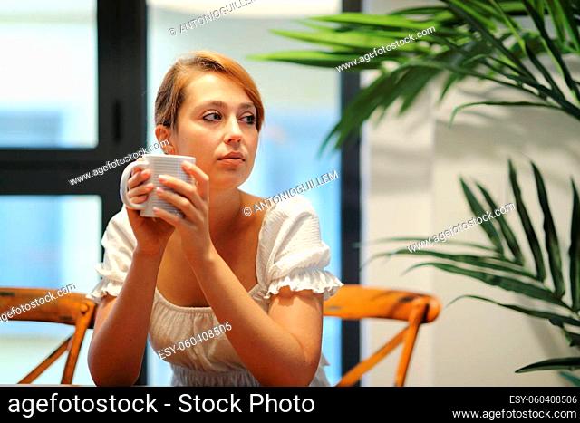 Serious woman drinking coffee thinking looking at side sitting on a chair at home