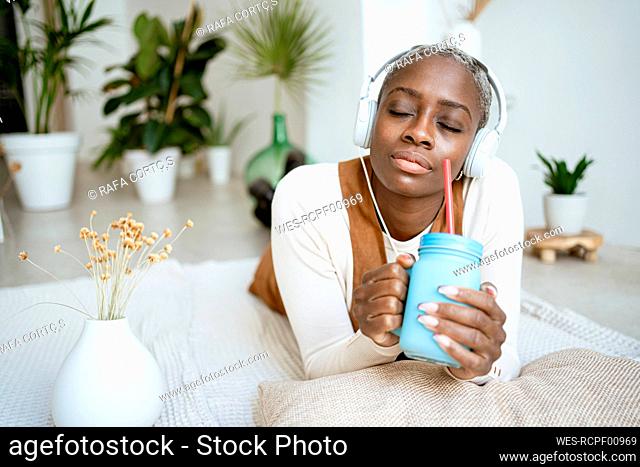 Woman with headphones holding jar while lying on rug in living room