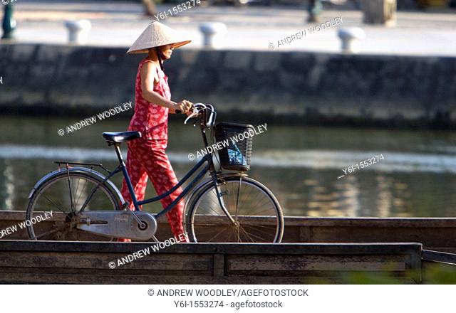 Conical hat woman pushes bicycle over footbridge Hoi An Vietnam