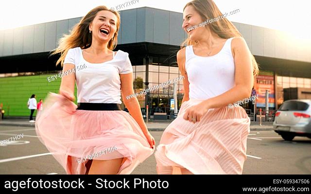 Portrait o two cheerful laughing girls in pink skirt running and jumping on car parking at shopping mall