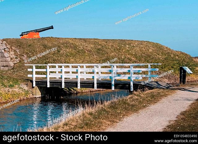 A wooden bridge over a small river. A gun on a hill. Open-air museum
