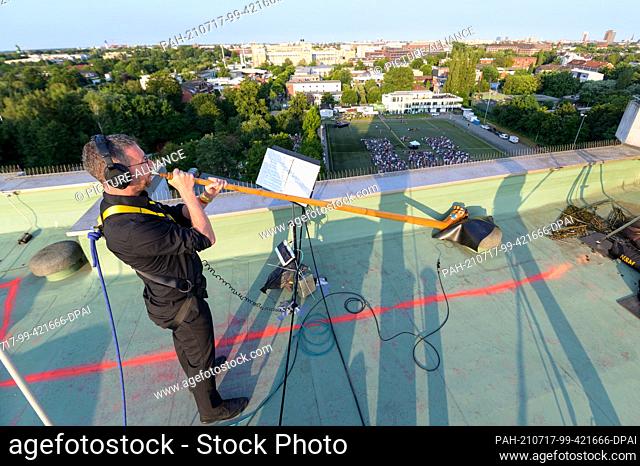 17 July 2021, Hamburg: A musician blows his alphorn on the roof of a high-rise building, while hundreds of spectators listen on a football pitch and all around