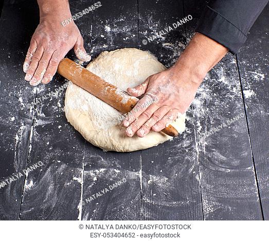 chef in a black tunic rolls a dough for a round pizza, top view
