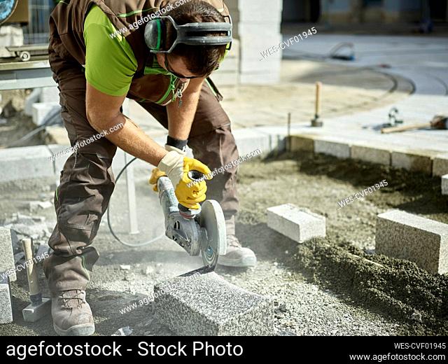 Blue-collar worker cutting paving stone by electric saw at construction site