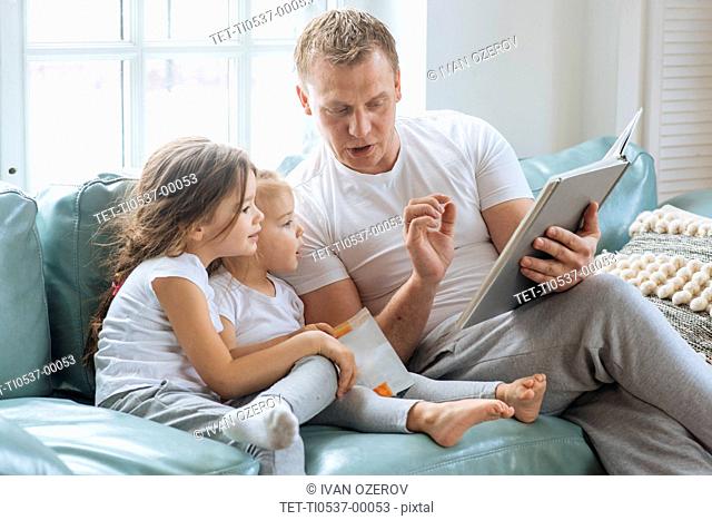 Father reading book to daughters on sofa