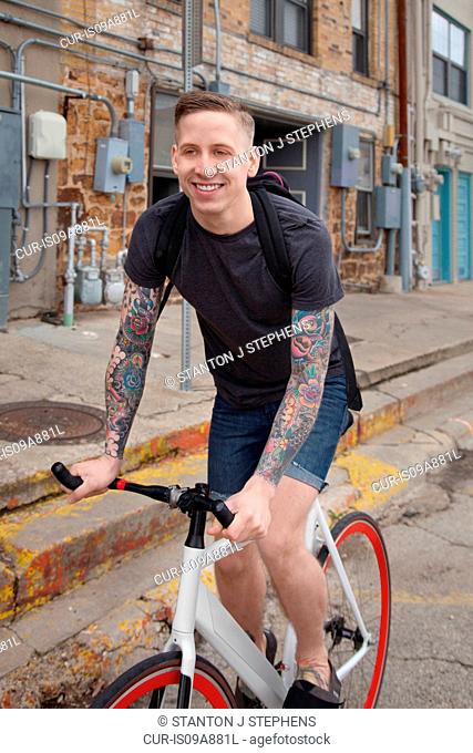 Young tattooed man cycling on street, smiling