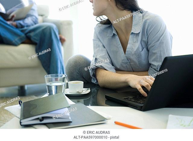 Woman using laptop in living room, looking over her shoulder at man sitting in background
