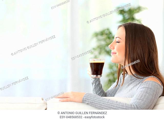 Profile portrait of a happy woman smelling a cup of coffee sitting on a couch at home