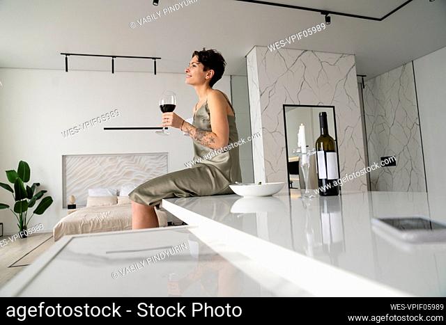 Smiling woman holding wineglass sitting on kitchen island at home