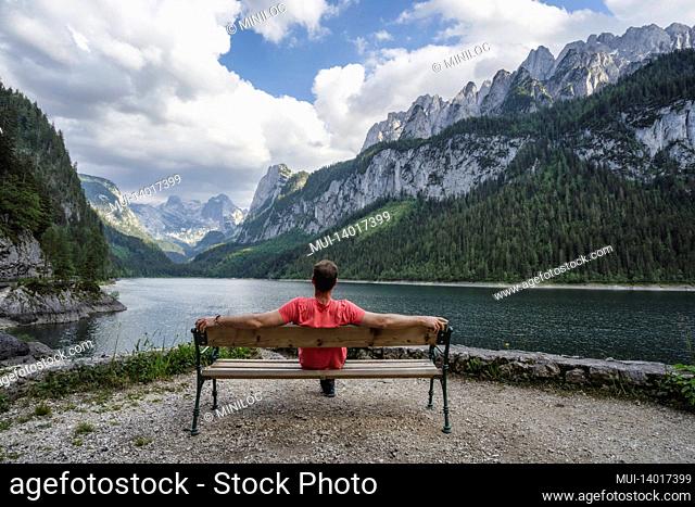 man relaxing on bench in front of dachstein mountains reflected in gosau lake, austria