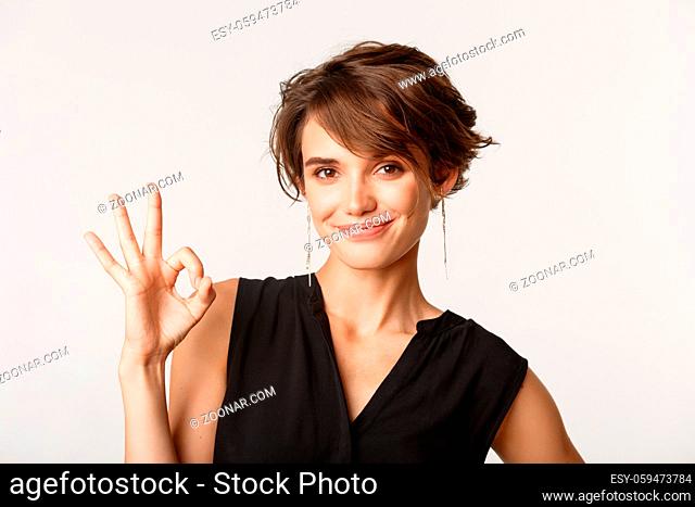 Close-up of confident businesswoman showing okay gesture and smiling satisfied, standing over white background