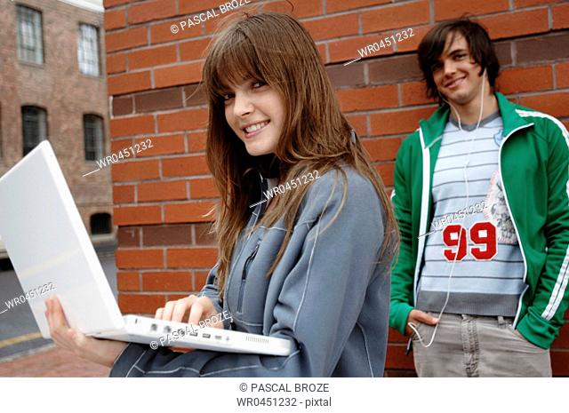 Portrait of a young woman using a laptop with a young man listening to music behind her