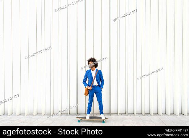 Happy businessman with shoulder bag standing on skateboard in front of wall