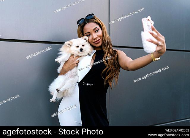 Young woman with dog taking selfie while standing in front of wall
