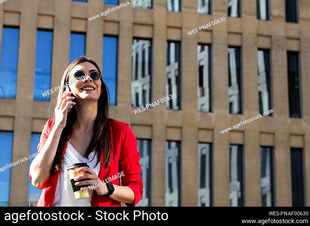 Smiling businesswoman talking on mobile phone while holding coffee cup against building