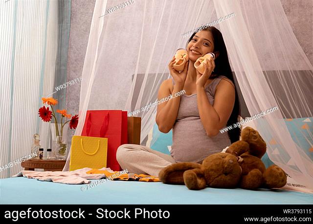Smiling pregnant woman holding a pair of baby shoes while sitting on bed at home